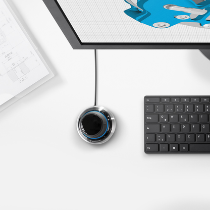 Top view of the 3Dconnexion SpaceMouse® Compact on a desk next to a keyboard and monitor displaying a 3D model, emphasizing precision.